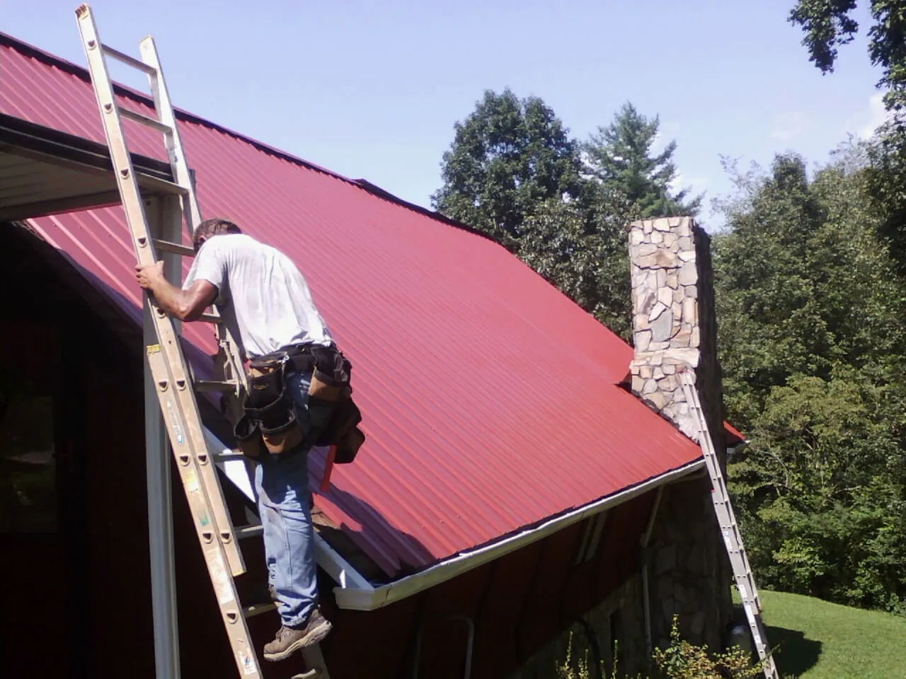 Roofer on a ladder beside a red metal roof and stone chimney — Soffit & Fascia Repair by Roofing Brothers in Atglen